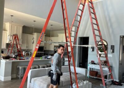 A person stands between two ladders set up in a living room with vaulted ceilings. There's another ladder and furniture, including a sofa and a rug, around the space.