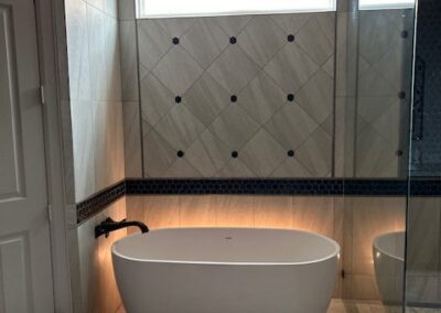 A modern bathroom features a white freestanding bathtub against a tiled wall, with a small window above. The floor is tiled, and a glass shower enclosure is visible on the right.