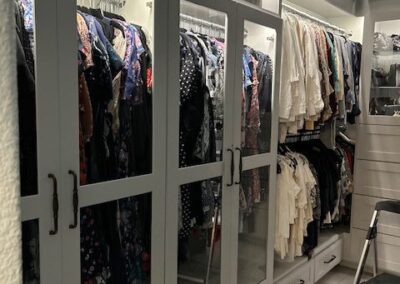 A walk-in closet with glass-door wardrobes, displaying neatly hung clothes in various colors and patterns. A step stool is placed nearby on the tiled floor.