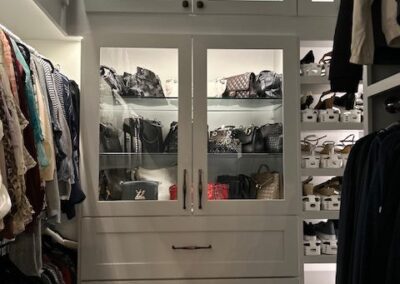 A well-organized walk-in closet with clothes hung on the left, shoes on shelves to the right, and handbags displayed behind glass doors in the center.