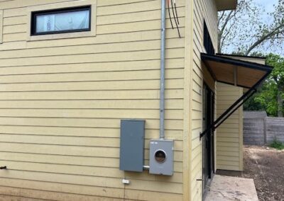 Side view of a beige building with a small window, an electrical meter box, a narrow roof overhang, and a dirt ground. A cable runs vertically along the wall.