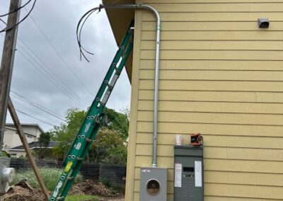 A green ladder leans against a yellow house. Electrical conduits and boxes are attached to the exterior wall near utility wires.