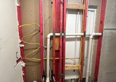 Exposed bathroom wall with visible plumbing and electrical wiring. Red framing, pipes, and yellow cables are present. Tools and construction materials are on the floor.