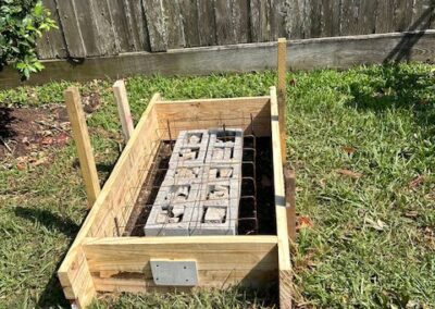 A wooden rectangular garden bed frame with cinder blocks inside is positioned on a grassy area near a weathered wooden fence.