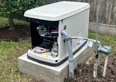 A generator on a concrete slab with its panel open, showing wires and components. Nearby, pipes and cables are connected. The setting is outdoors, with grass and a wooden fence in the background.