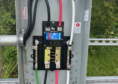 A metal electrical panel box with exposed wiring and various colored cables, surrounded by greenery outside.