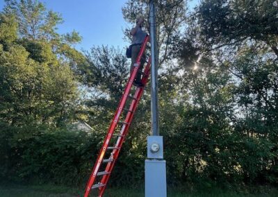 A person on a tall red ladder works on a utility pole surrounded by trees under a clear blue sky. A box and some items are on the ground nearby.