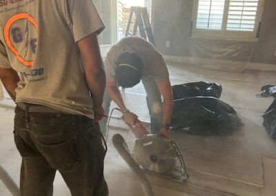 Two workers in a dusty room are using a large circular saw. One operates the saw while the other stands nearby. A ladder is visible in the background near a window with shutters.