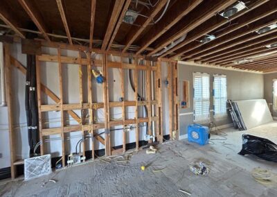 Room under renovation with exposed wooden beams, wiring, and pipes. A blue dehumidifier is on the floor, and construction materials are scattered around.